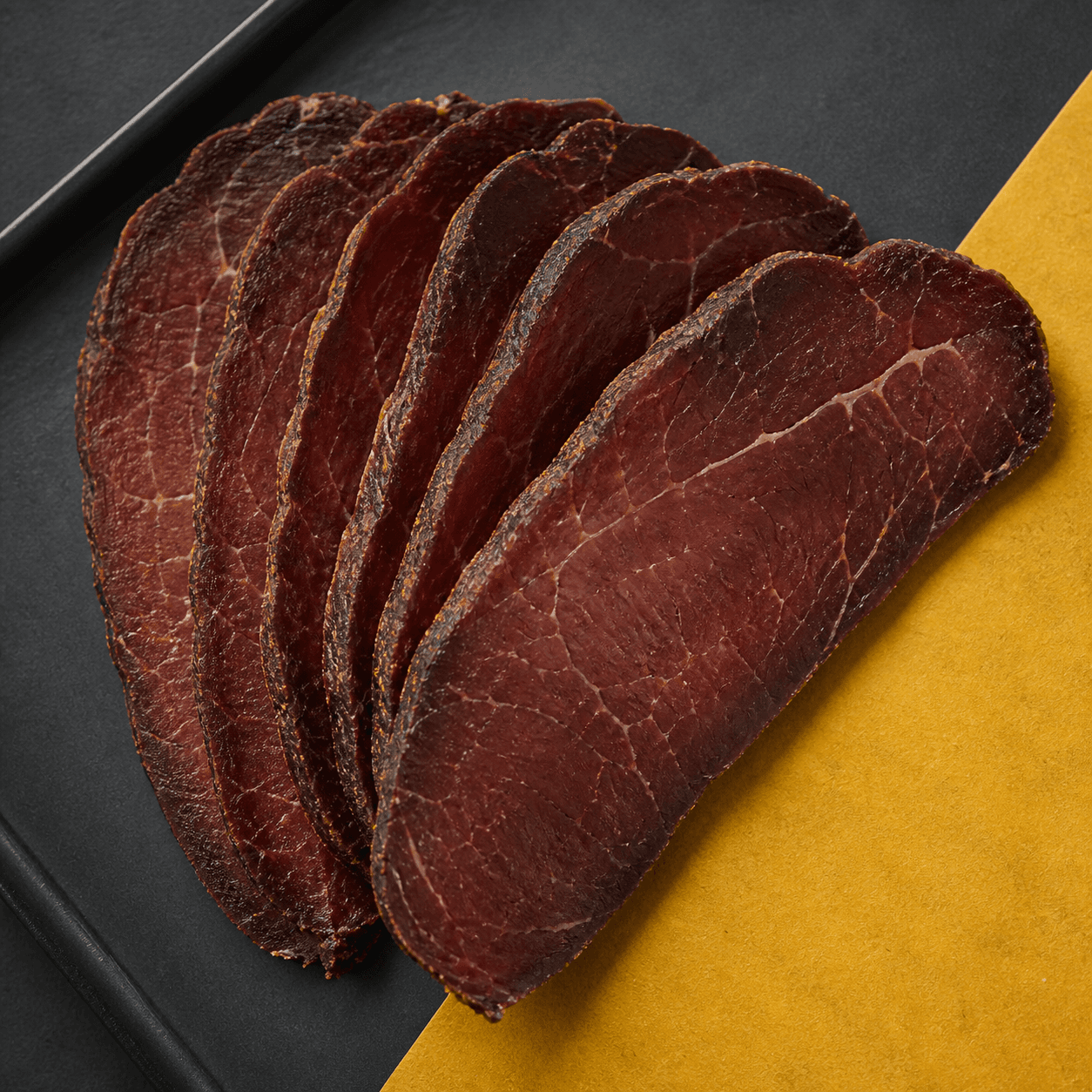 Thick slices of dark air-dried beef fanned across a charcoal tray and yellow paper