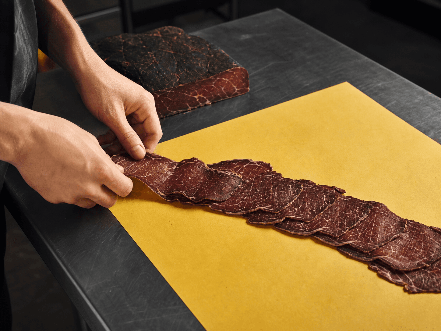 Hands arranging slices of air-dried beef on yellow paper across a charcoal preparation counter